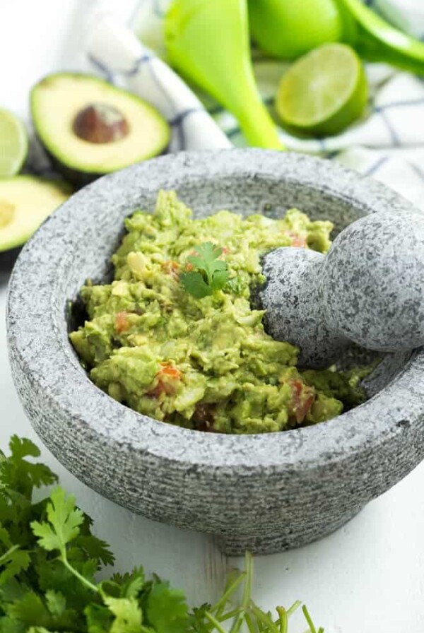 guacamole in a mortary with a spring of cilantro on top. Avocado, cilantro and lemon on the table next to the bowl.