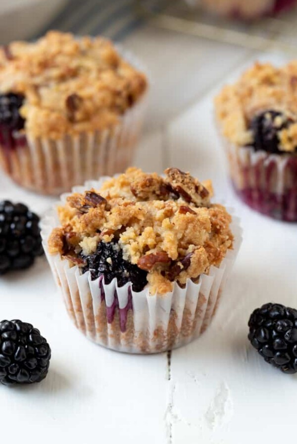 three blackberry muffins sitting on a white table next to a few fresh blackberries