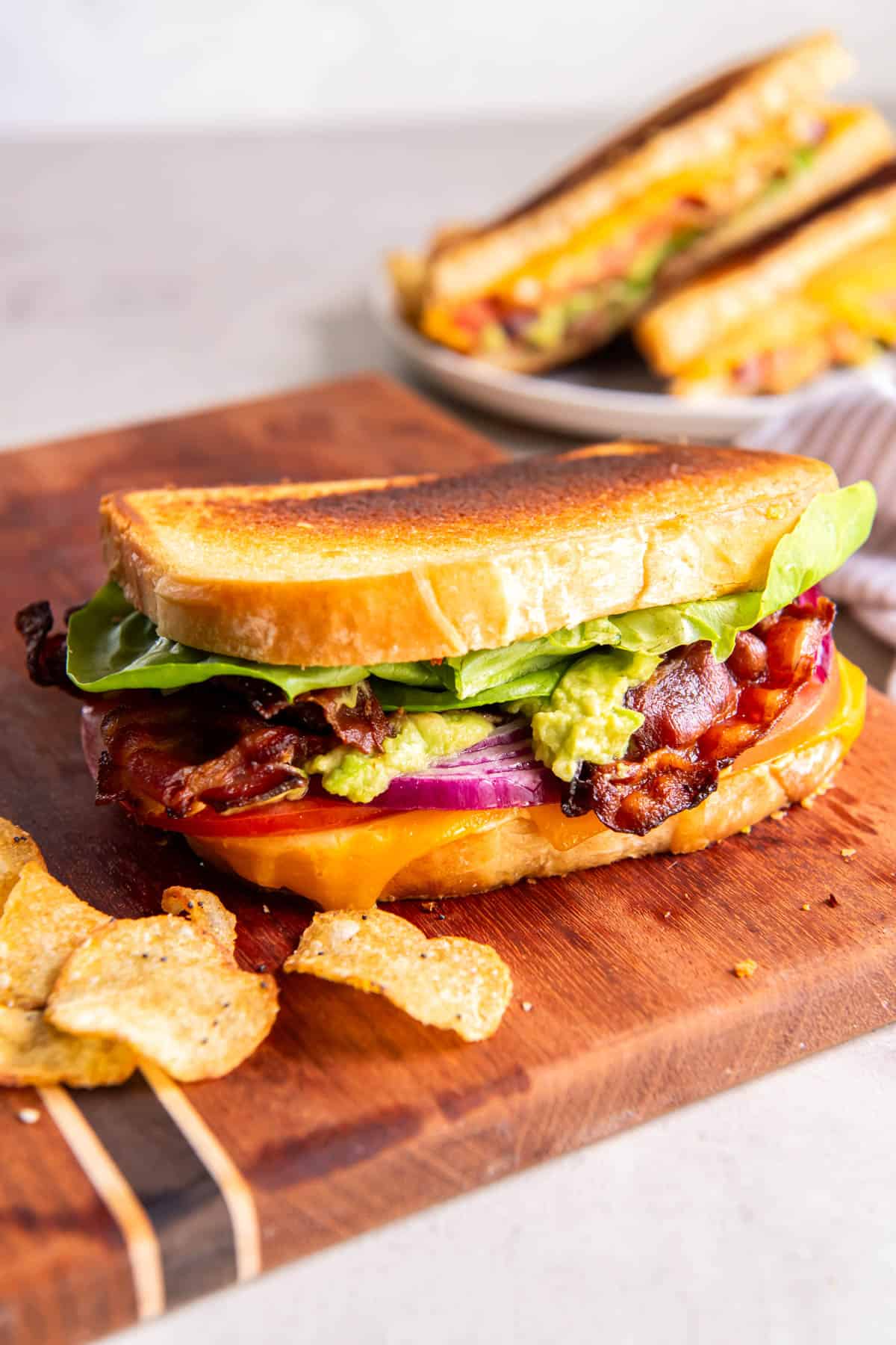 BLT grilled cheese sitting on a cutting board with a side of chips. Another sandwich sits on a plate on the back of the cutting board. 