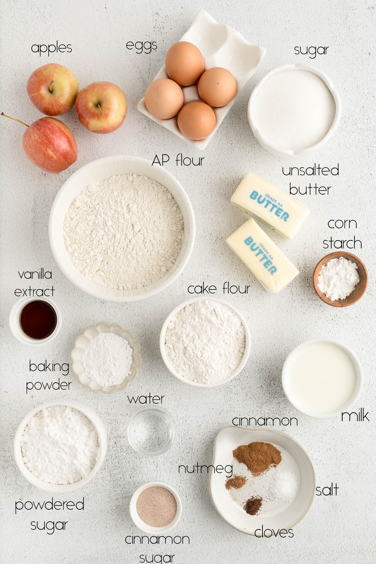 All of the ingredients for apple pie cupcakes in white bowls on a white countertop. 