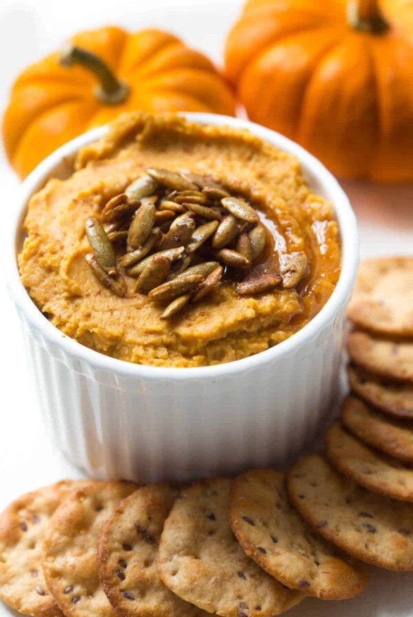 a white bowl with pumpkin hummus sitting next to a stack of crackers and two mini pumpkins