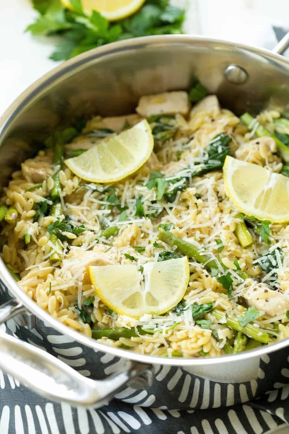 A Pan of Lemon Chicken Orzo with spinach, asparagus, parmesan cheese, and lemon wedges.