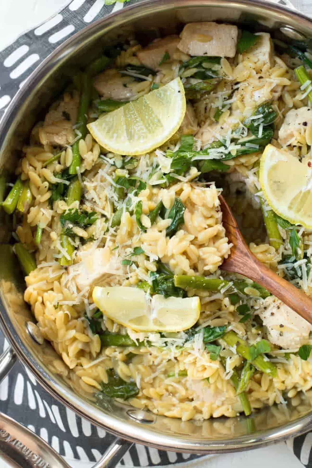 A Pan of Lemon Chicken Orzo with spinach, asparagus, and lemon wedges. A spoon is scooping some out