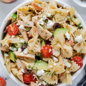 Greek chicken pasta salad in a white bowl for serving. Cucumbers, tomatoes, kalamata olives, cooked chicken, bowtie pasta and feta cheese are piled high in the bowl.