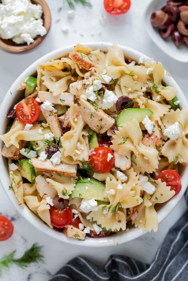 Greek chicken pasta salad in a white bowl for serving. Cucumbers, tomatoes, kalamata olives, cooked chicken, bowtie pasta and feta cheese are piled high in the bowl.