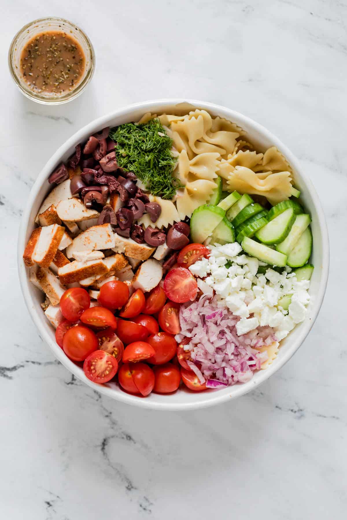 Ingredients for the greek chicken salad sit in a white bowl before mixing including cooked bow tie pasta, cherry tomatoes, cucumber, kalamata olives, red onion, feta cheese and fresh dill. A small glass with the homemade vinaigrette sits on the side.