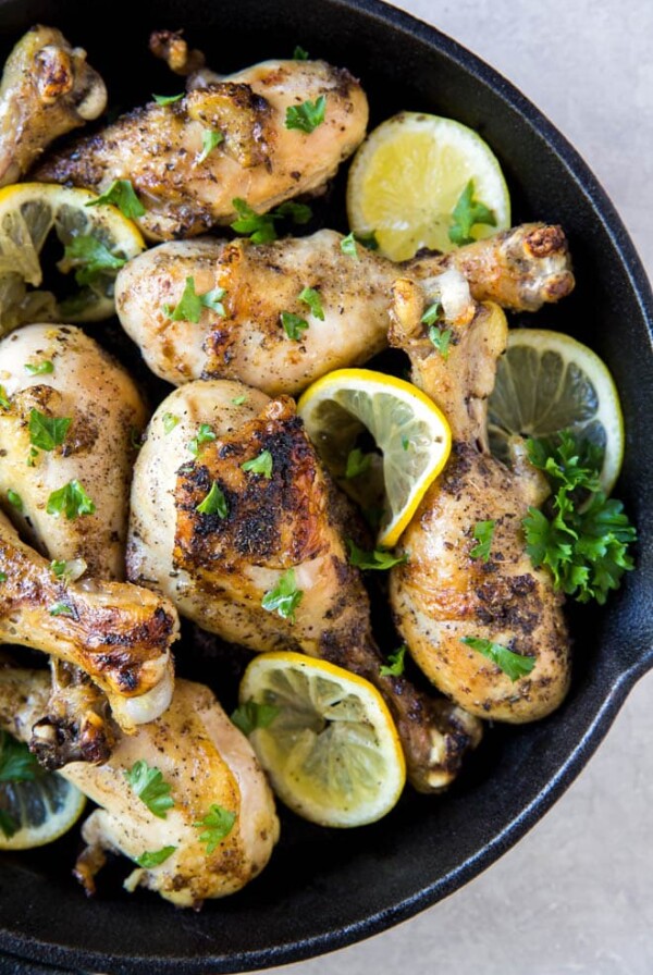 baked lemon pepper chicken drumsticks layered in a cast iron skillet with parsley garnish and slices of lemons