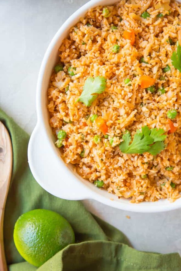 A large bowl of Mexican cauliflower rice sitting on a table next to a green napkin and a lime.