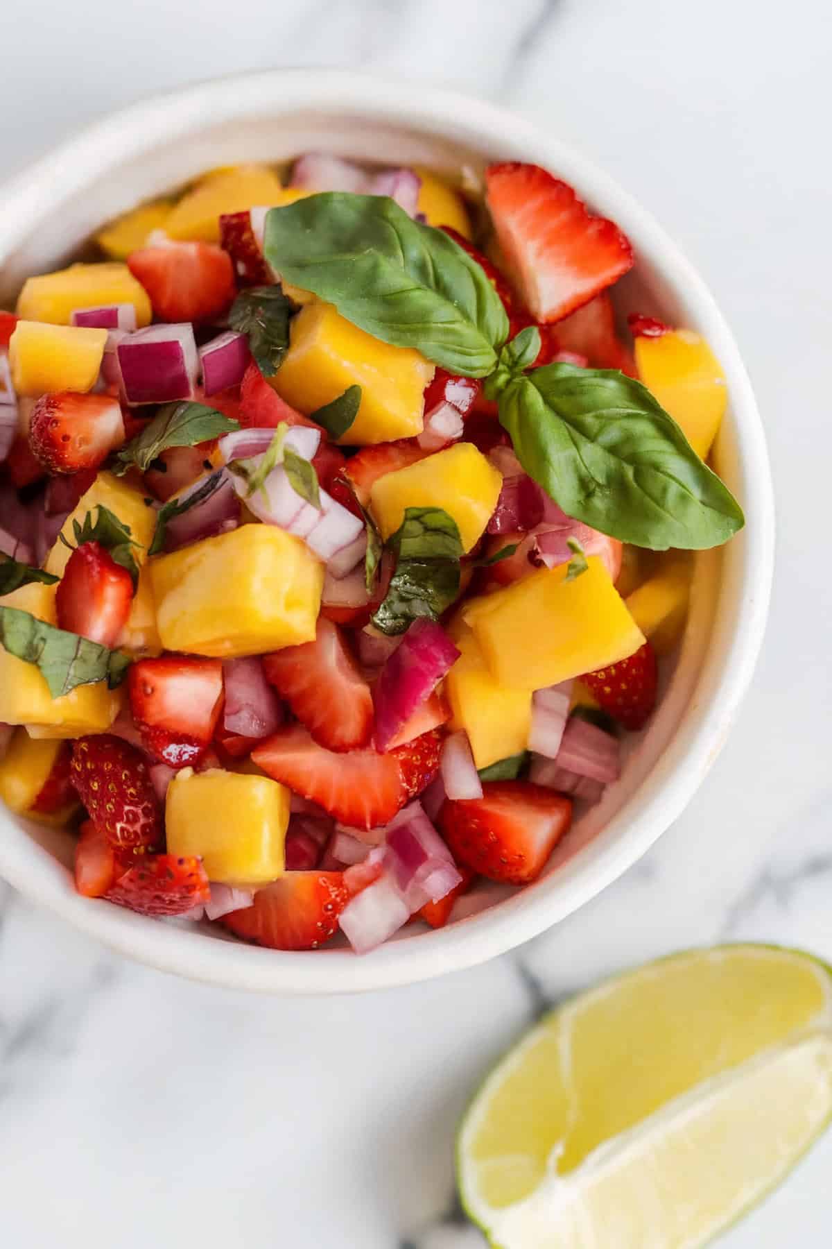 An overhead shot of a bowl of strawberry mango salsa sitting on a white background with a lime wedge on the side.