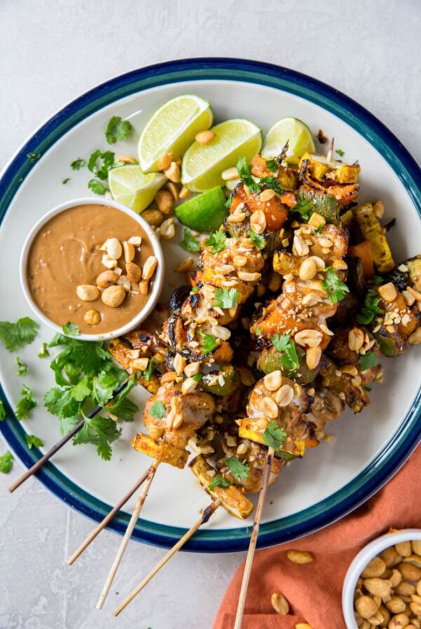 a pile of chicken kebabs sitting on a round plate with a bowl of peanut sauce and sliced limes on the side.