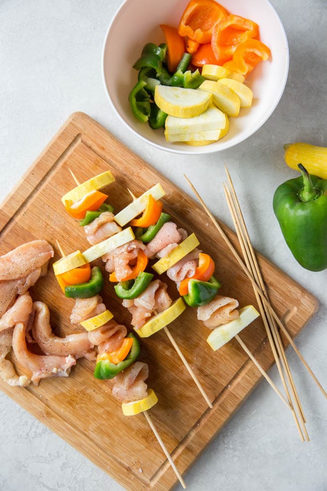 Chicken kebabs sit on skewers on a wood cutting board before grilling.