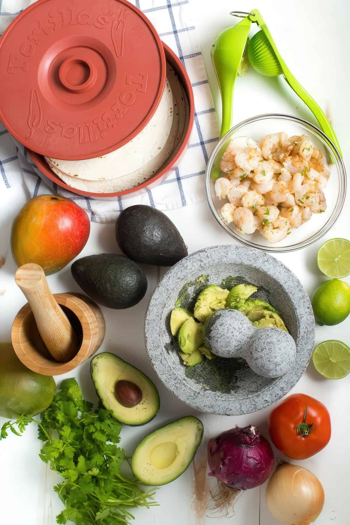 A top-down view of fresh ingredients for shrimp tacos with mango avocado salsa. The spread includes a tortilla warmer filled with soft tortillas, a bowl of seasoned shrimp, ripe avocados, a mortar and pestle with mashed avocado, a wooden pestle and mortar, a mango, limes, a green citrus juicer, fresh cilantro, a red onion, a white onion, and a tomato, all arranged on a white surface with a checkered cloth.