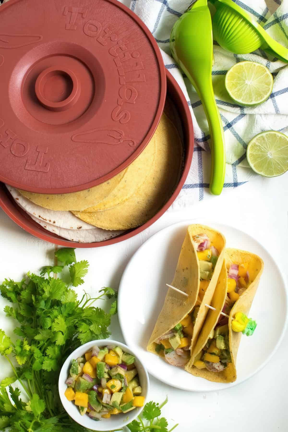 Shrimp tacos with mango avocado salsa on a white plate, accompanied by a small bowl of salsa, fresh cilantro, and a tortilla warmer filled with corn and flour tortillas. A green citrus juicer and halved limes rest on a checkered cloth in the background. The tacos are held together with wooden toothpicks, one decorated with a small green and yellow paper umbrella. 