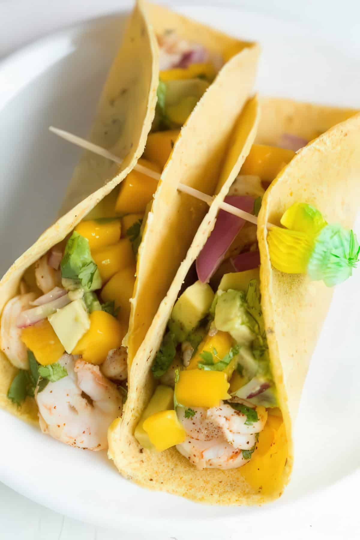 three shrimp tacos on a white plate, filled with seasoned shrimp, diced mango, avocado, red onion, and cilantro in soft corn tortillas. The tacos are held together with wooden toothpicks, one of which is decorated with a small green and yellow paper umbrella.