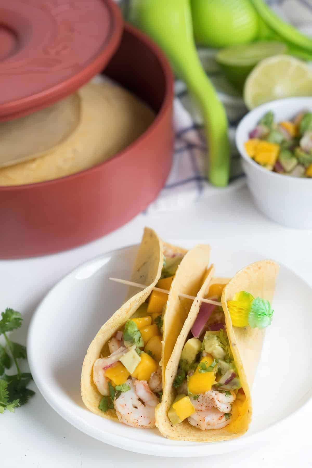 A close-up of three shrimp tacos served on a white plate, filled with shrimp, diced mango, avocado, red onion, and cilantro in soft corn tortillas. The tacos are held together with decorative toothpicks featuring small paper umbrellas. In the background, a red tortilla warmer filled with tortillas, a bowl of mango avocado salsa, fresh limes, and a green citrus juicer.