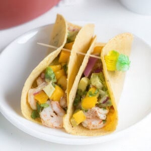 Three shrimp tacos on a white plate, filled with seasoned shrimp, diced mango, avocado, red onion, and cilantro in soft corn tortillas. The tacos are held together with wooden toothpicks, one of which is decorated with a small green and yellow paper umbrella.