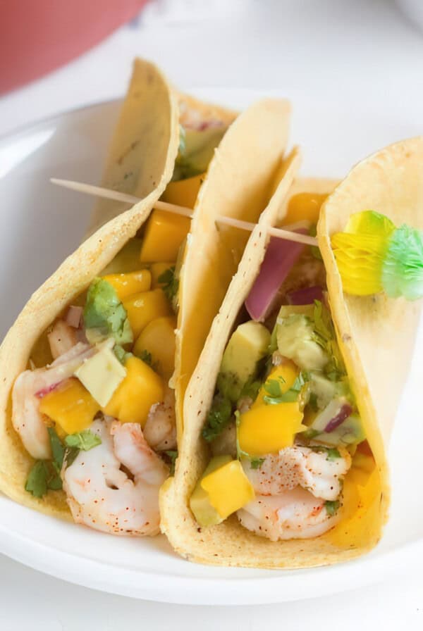 Three shrimp tacos on a white plate, filled with seasoned shrimp, diced mango, avocado, red onion, and cilantro in soft corn tortillas. The tacos are held together with wooden toothpicks, one of which is decorated with a small green and yellow paper umbrella.