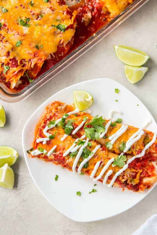 Two turkey enchiladas sit on a white plate with a casserole dish full of enchiladas off to the side.