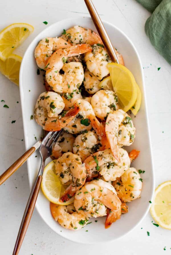 Cooked lemon garlic shrimp on a white serving plate with serving fork and lemon wedges.