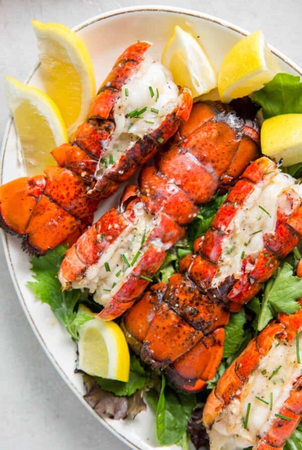 Four grilled lobster tails on a bed of lettuce on a plate with lemon wedges.