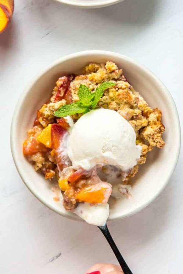 A large bowl of peach crumble with a scoop of vanilla ice cream and a spoon.