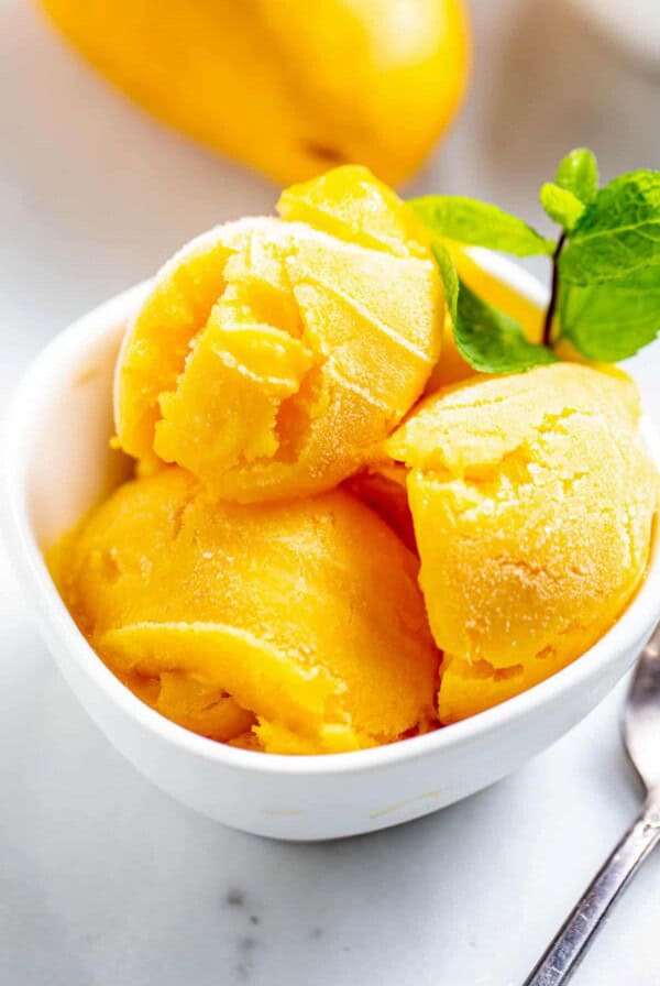 Three scoops of homemade mango sorbet in a white bowl with a sprig of peppermint on top.