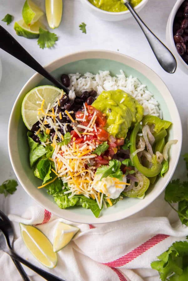 A white bowl filled with white rice, sauteed peppers and onions, guacamole, black beans, lettuce, pico de gallo and shredded cheese. A fork sits in the bowl.