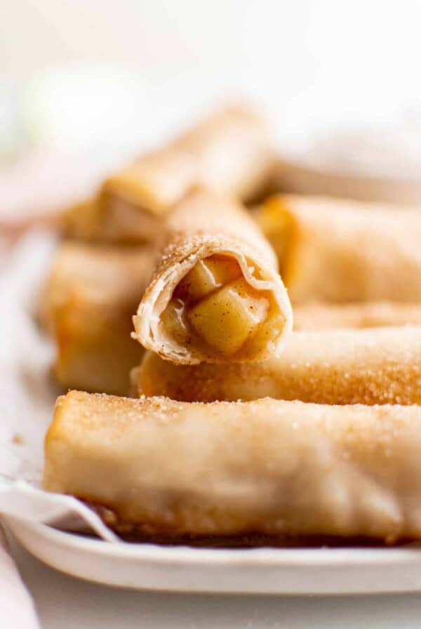 One apple pie egg roll with a bite taken out of it sitting on top of more apple pie egg rolls on a baking sheet.