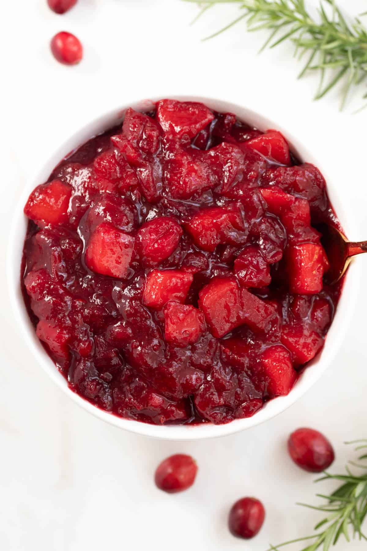 apple cranberry sauce in a white bowl on a white countertop with some fresh rosemary and whole cranberries on the side for garnish.