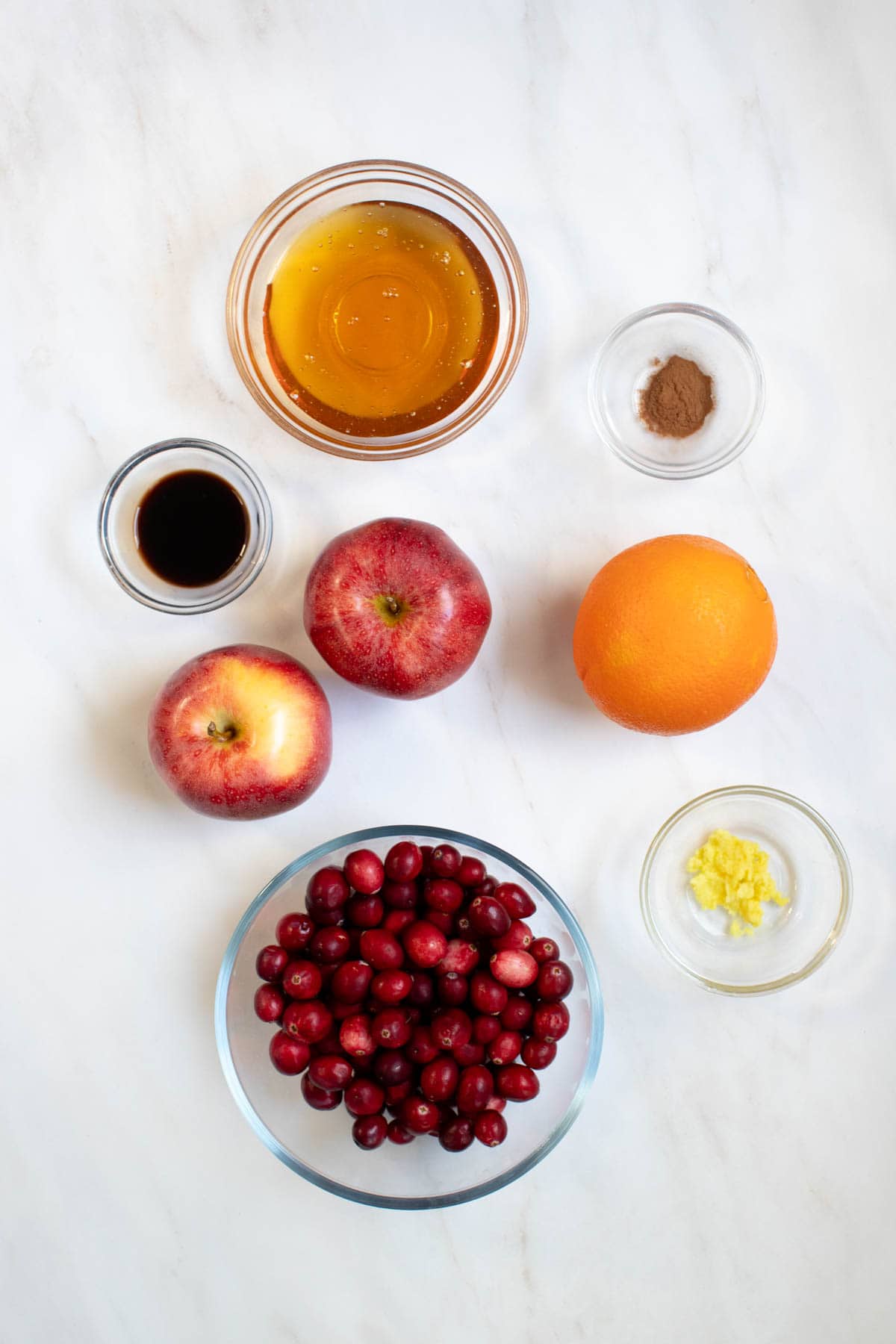 All of the ingredients for cranberry apple sauce in bowls on a white countertop. 