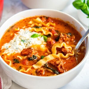 A white bowl filled with lasagna soup sits on a white countertop. A spoon sits in the bowl for serving.