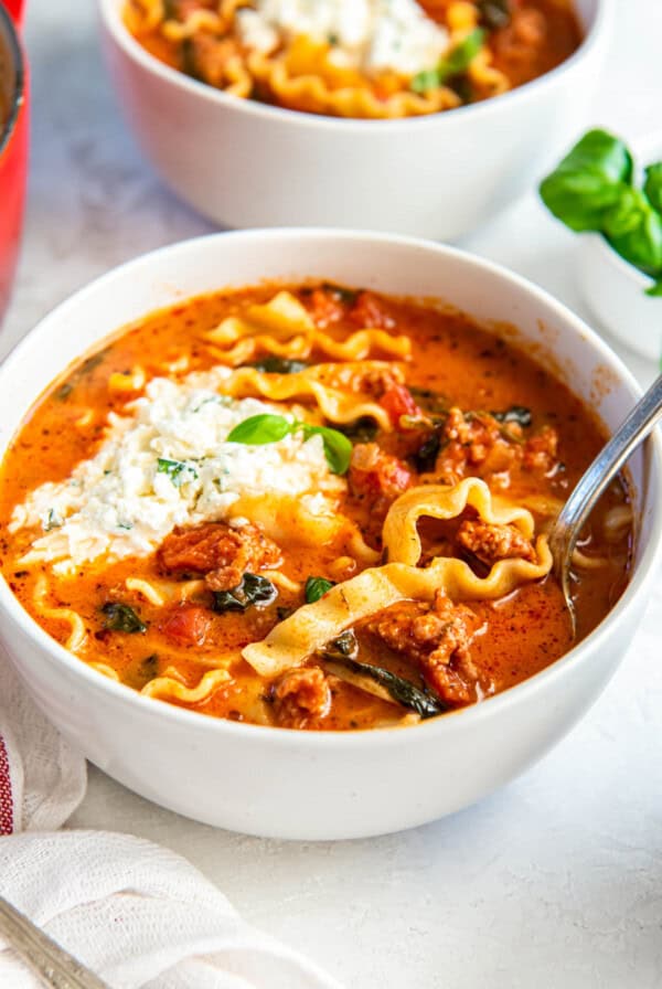 A white bowl filled with lasagna soup sits on a white countertop. A spoon sits in the bowl for serving.