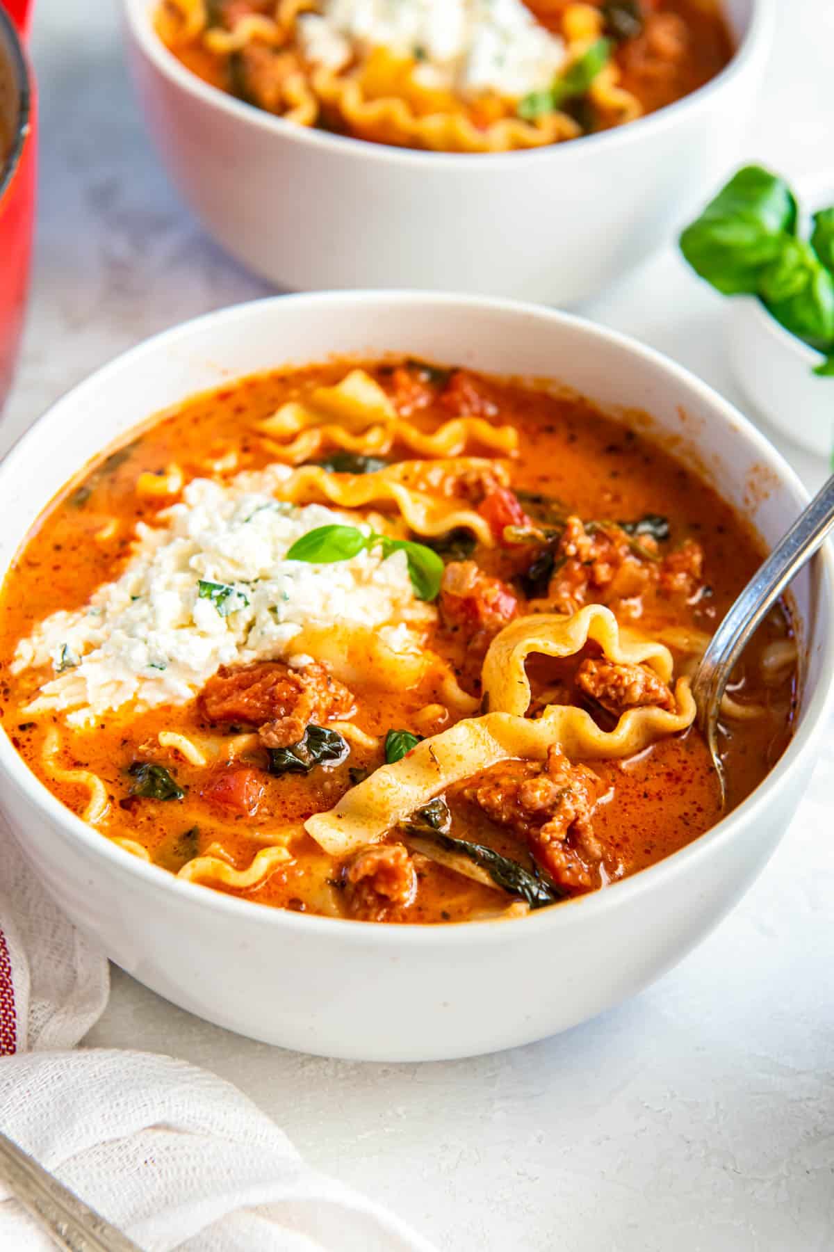 A white bowl filled with lasagna soup sits on a white countertop. A spoon sits in the bowl for serving. 