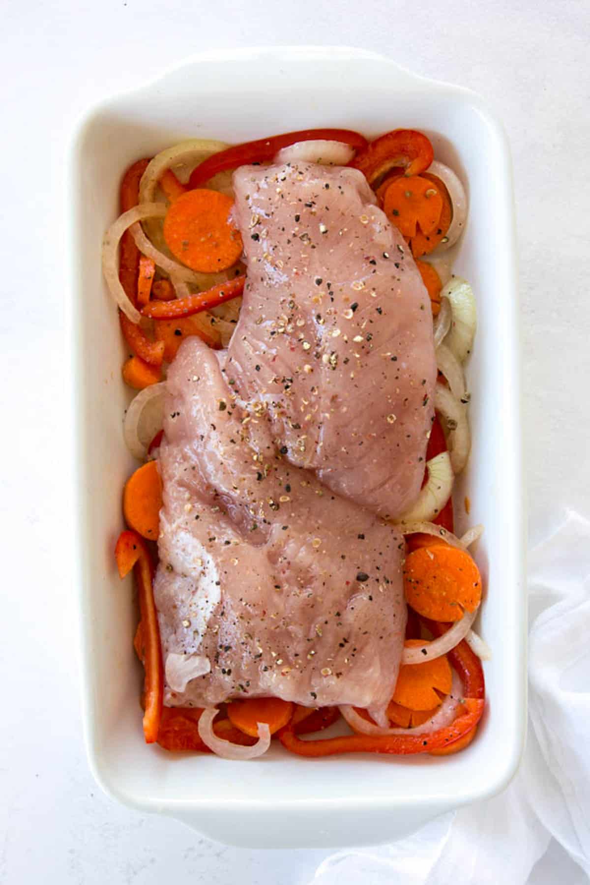 Sliced carrots, onions and red bell peppers in a white dish with a turkey tenderloin on top of the vegetables before baking. 