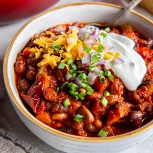 A bowl filled with three bean chili and topped with sour cream, cheddar cheese, red onion and chopped chives. A spoon also sits in the bowl for serving.