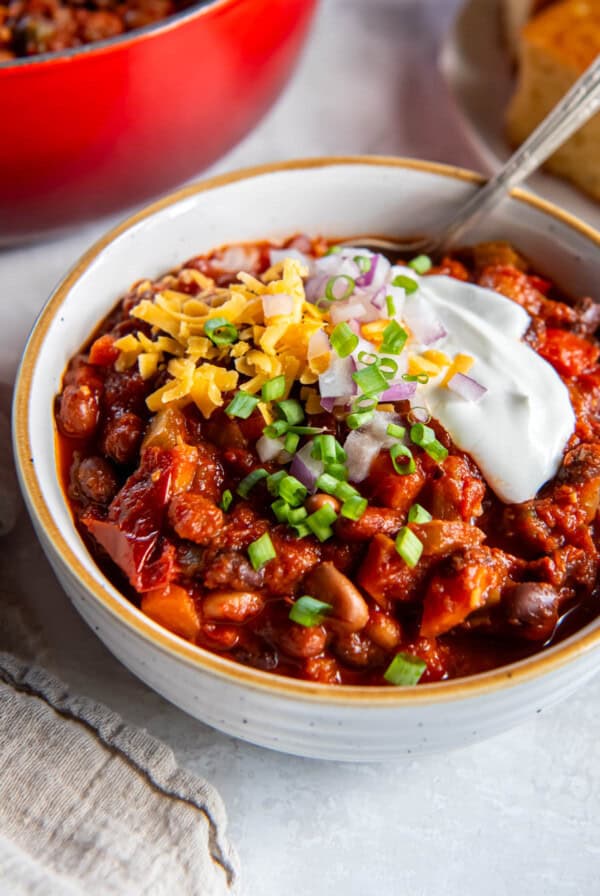 A bowl filled with three bean chili and topped with sour cream, cheddar cheese, red onion and chopped chives. A spoon also sits in the bowl for serving.