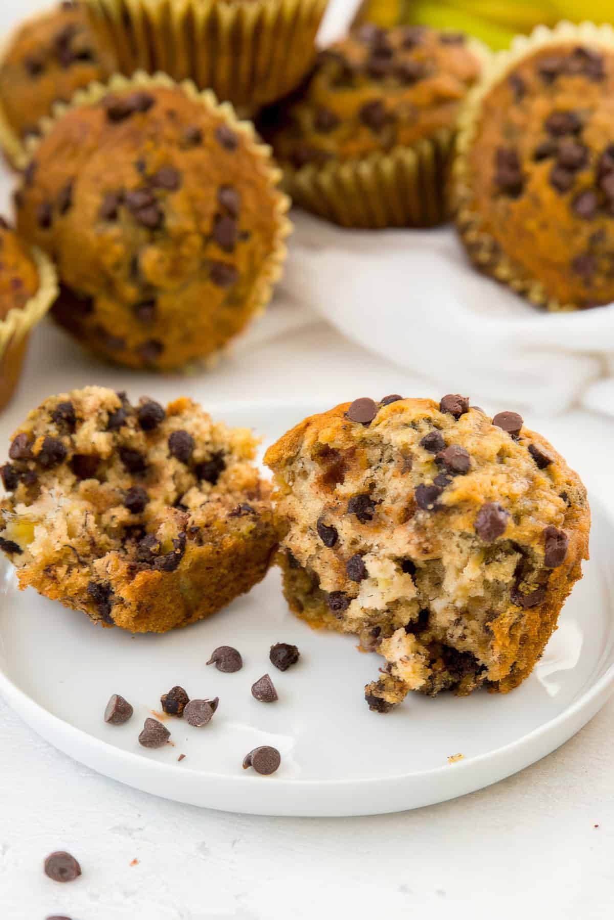 a banana chocolate chip muffin broken in half and sitting on a white plate with other muffins in the background.