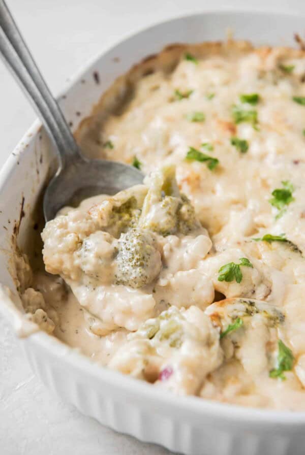 Cooked broccoli cauliflower casserole in a white baking dish on a white countertop. A serving spoon is scooping some of the casserole.