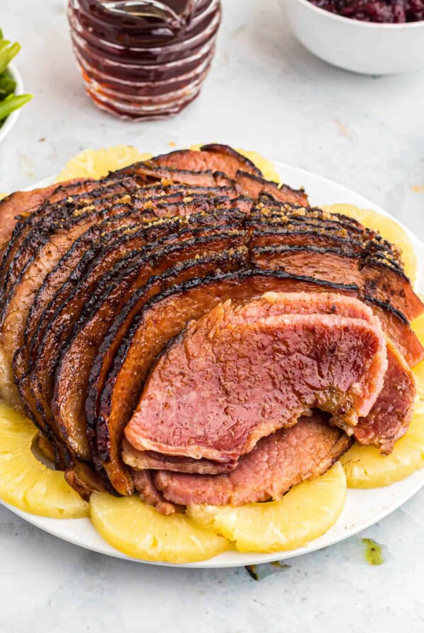 a cooked maple glazed ham sitting on a white serving platter with pineapple around the edges