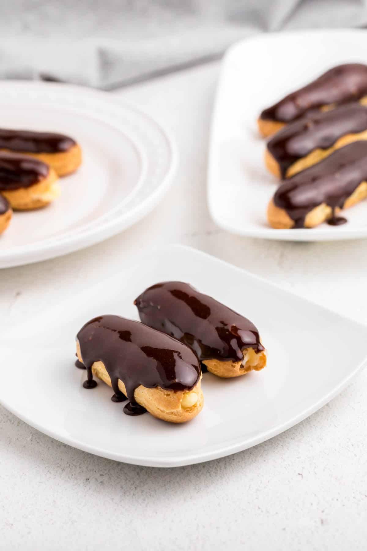 Two freshly baked eclairs topped with a chocolate glaze on a square white plate.