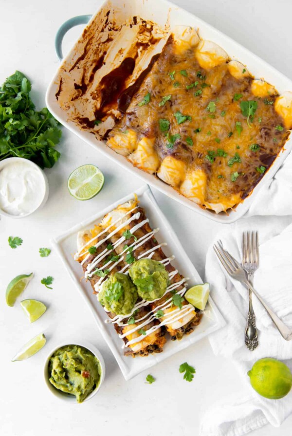 Aerial view of a pan of veggie enchiladas and a plate with two vegetable enchiladas, topped with guacamole and sour cream.