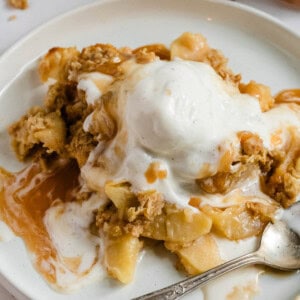 Delicious apple crispy with oatmeal topping sits on a white plate with a scoop of vanilla ice cream on top.