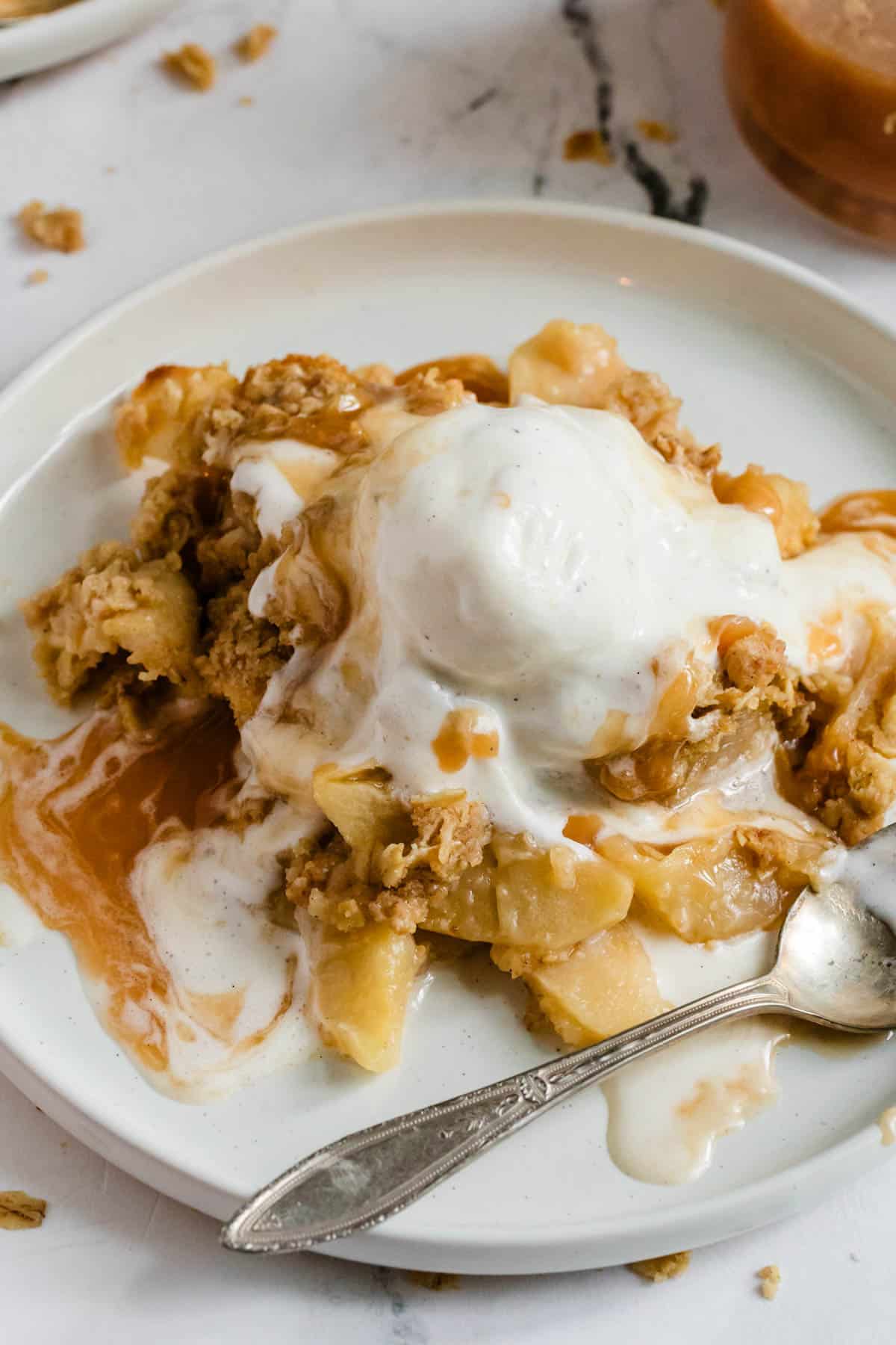 Delicious apple crispy with oatmeal topping sits on a white plate with a scoop of vanilla ice cream on top.