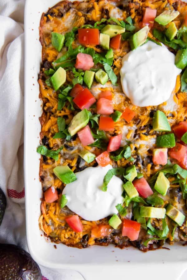 A white casserole dish filled with taco casserole and topped with chopped tomato, avocado, cilantro and sour cream.