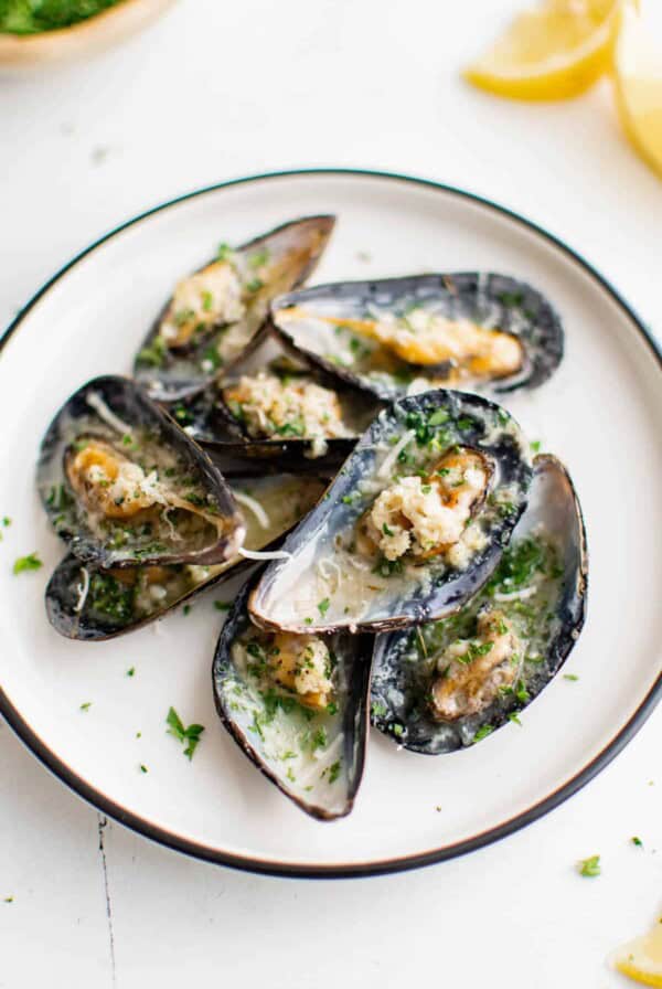 Eight baked mussels in garlic butter on a white plate sitting on a white tabletop for serving. They are garnished with freshly chopped parsley.