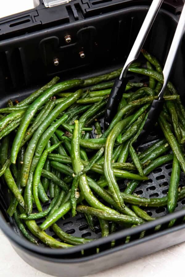 Tongs serving cooked air fryer green beans out of the air fryer basket.