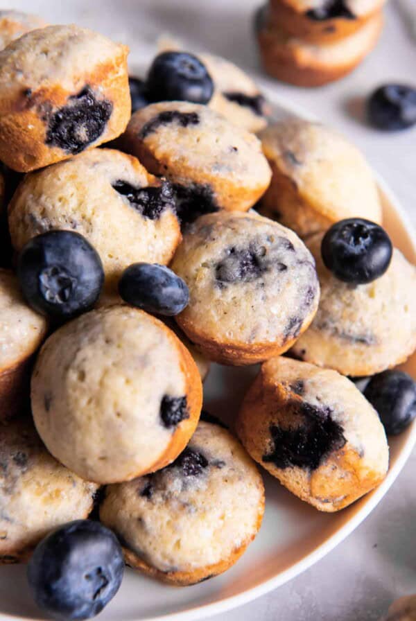 Mini blueberry muffins sitting on a plate for serving. Fresh blueberries are scattered on top of the muffins.