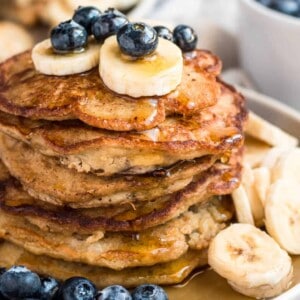 A stack of banana oatmeal pancakes on a plate served with fresh blueberries and bananas and drizzled with maple syrup.