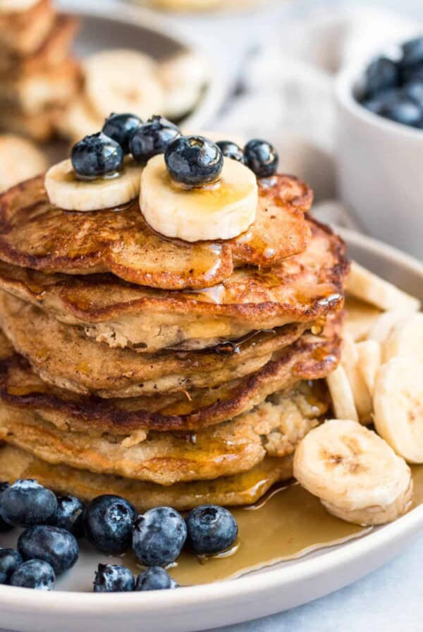 A stack of banana oatmeal pancakes on a plate served with fresh blueberries and bananas and drizzled with maple syrup.