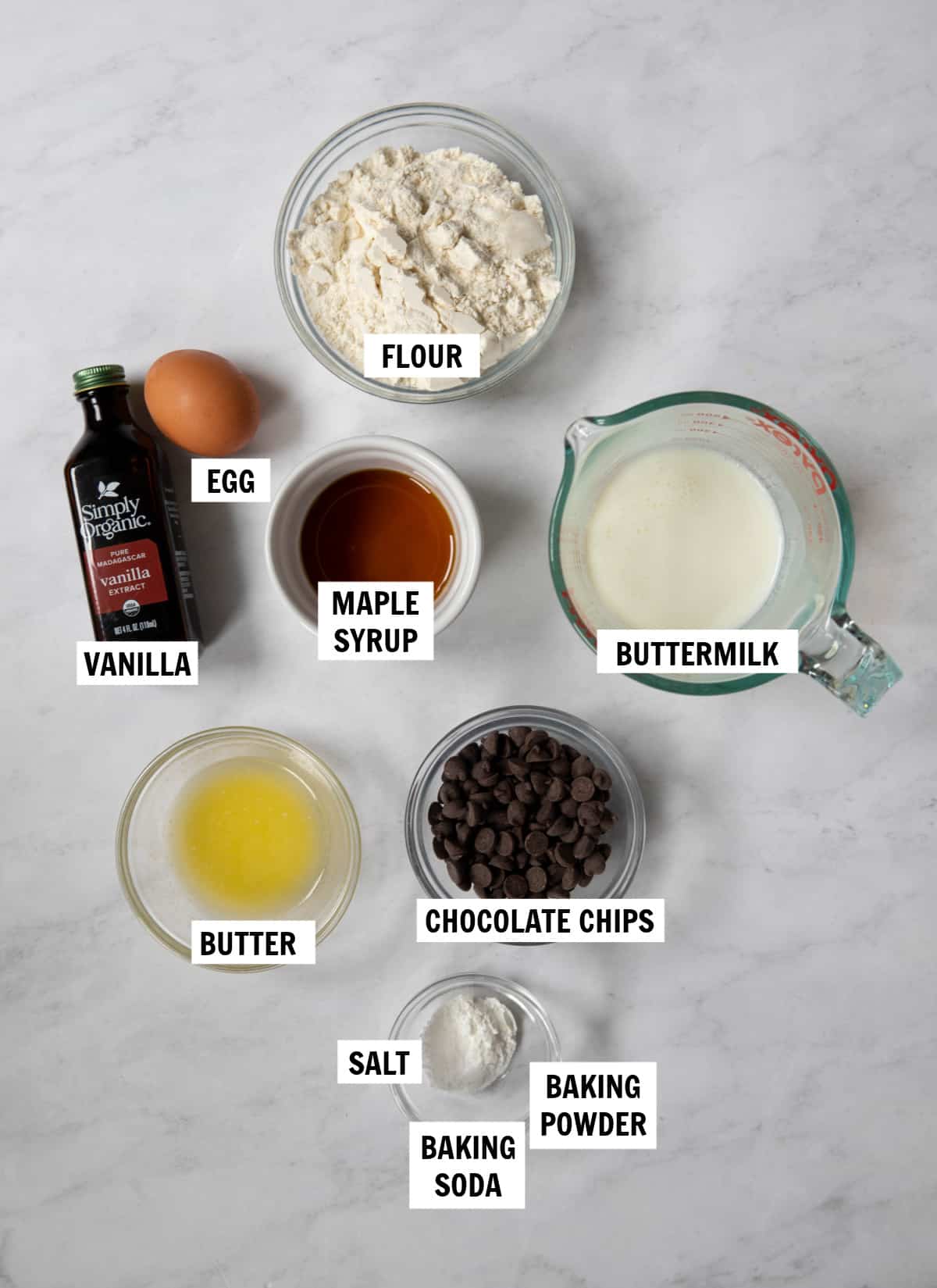 All of the ingredients for pancake bites on a white countertop in prep bowls including flour, buttermilk, egg, vanilla, maple syrup, butter, chocolate chips, salt, baking powder and baking soda.